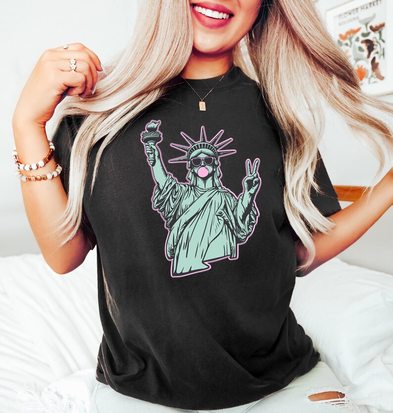Może przedstawiać: Czarny t-shirt z grafiką przedstawiającą Statuę Wolności w okularach przeciwsłonecznych i z bańką z gumy do żucia. Statua jest w kolorze jasnoniebieskim z r&oacute;żowym obrysem. Koszulka jest w ciemnym kolorze.