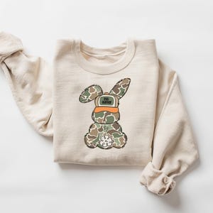 May include: A beige sweatshirt with a cartoon bunny wearing a camouflage pattern and a hat that says "Egg Hunter".