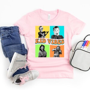 May include: Pink t-shirt with a colorful graphic of four kids from the movie Home Alone. The graphic includes the text "Kid Vibes".