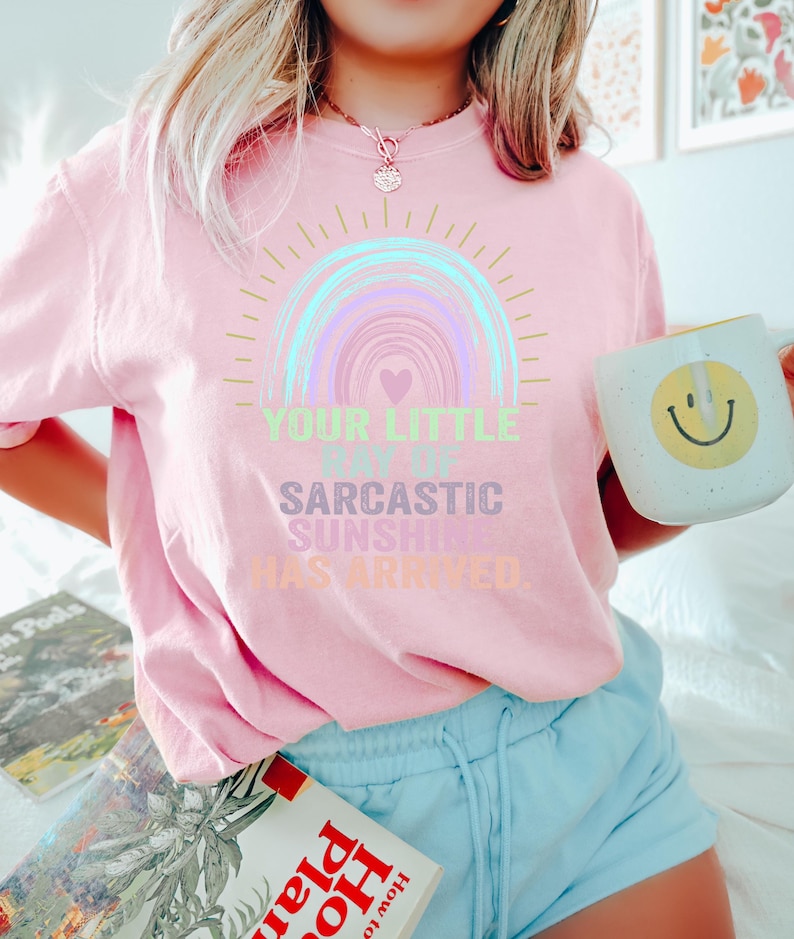 May include: A pink t-shirt with a rainbow graphic and the text "Your Little Ray of Sarcastic Sunshine Has Arrived."