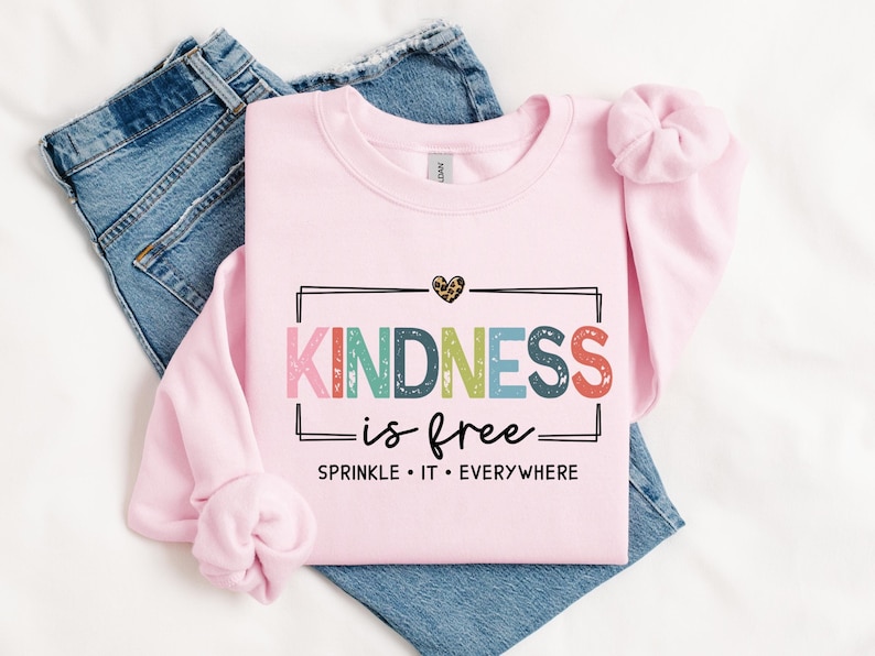 May include: Pink sweatshirt with the text "KINDNESS is free" in rainbow colors with a black border and a small leopard print heart above the text. The text "SPRINKLE IT EVERYWHERE" is below the main text.