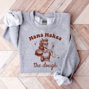 May include: Gray sweatshirt with a retro-style illustration of a woman holding a loaf of bread. The text "Mama Makes the Dough" is printed on the sweatshirt.
