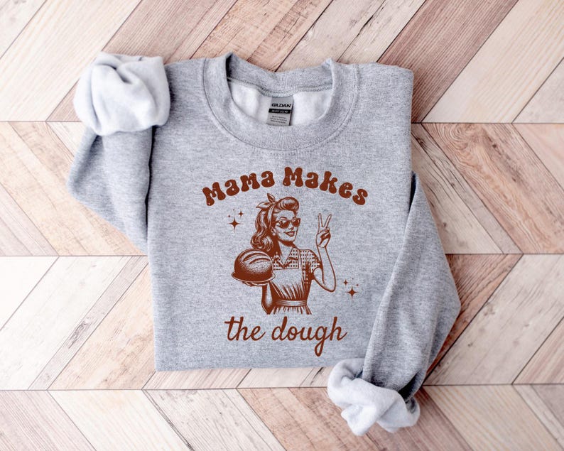May include: Gray sweatshirt with a retro-style illustration of a woman holding a loaf of bread. The text "Mama Makes the Dough" is printed on the sweatshirt.