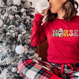 Puede incluir: Una camisa roja de manga larga con un gr&aacute;fico colorido que dice "NURSE" con un fantasma sosteniendo una flor. La camisa la lleva puesta una persona que lleva pantalones de pijama a cuadros.