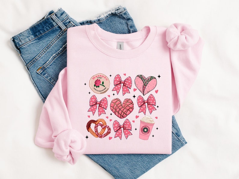May include: Pink sweatshirt with a graphic design featuring a variety of Mexican treats, including a heart-shaped concha, a heart-shaped taco, and a marzipan candy. The graphic also includes pink bows and stars.