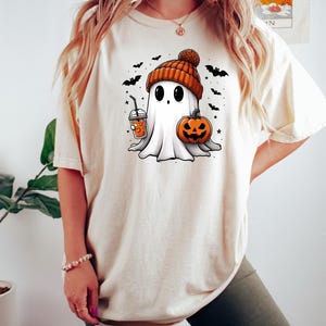 May include: A white t-shirt with a cartoon ghost wearing an orange and brown hat, holding a pumpkin and a cup of orange liquid. The ghost is surrounded by bats and stars.