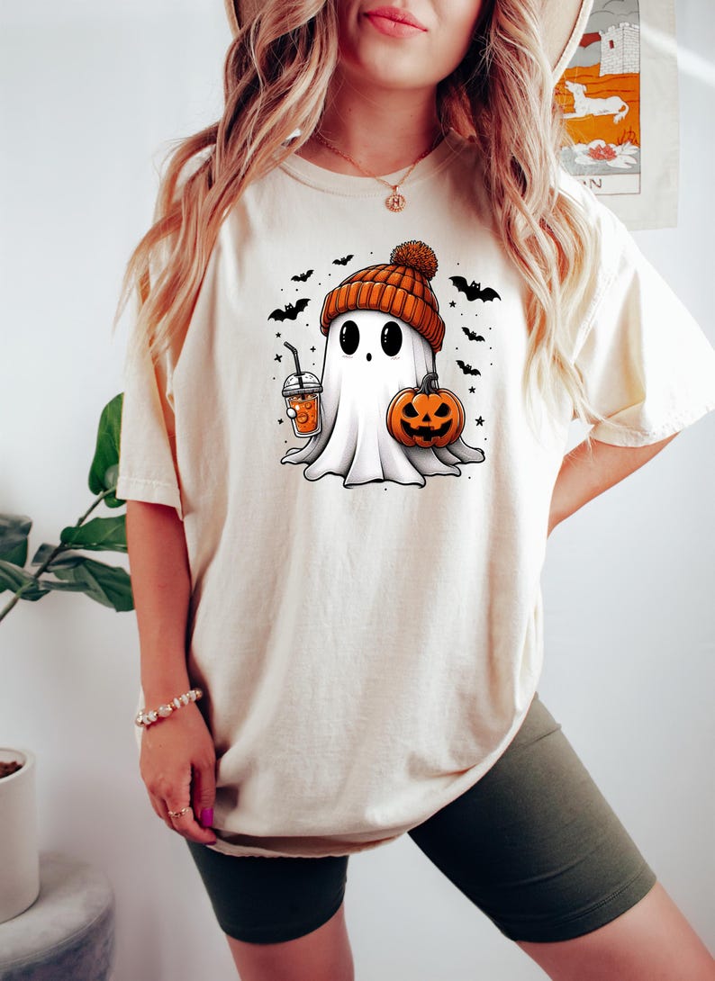 May include: A white t-shirt with a cartoon ghost wearing an orange and brown hat, holding a pumpkin and a cup of orange liquid. The ghost is surrounded by bats and stars.