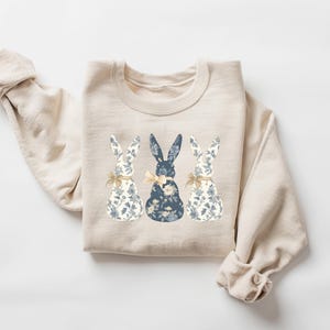 K&ouml;nnte beinhalten: Ein beiger Pullover mit einem floralen Muster aus drei blauen Hasen auf der Vorderseite.