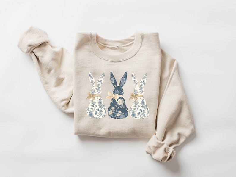 K&ouml;nnte beinhalten: Ein beiger Pullover mit einem floralen Muster aus drei blauen Hasen auf der Vorderseite.