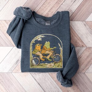 May include: A dark blue sweatshirt with a graphic of two frogs riding a bicycle. The frogs are green and brown and are wearing hats. The bicycle is red and has a basket on the front.
