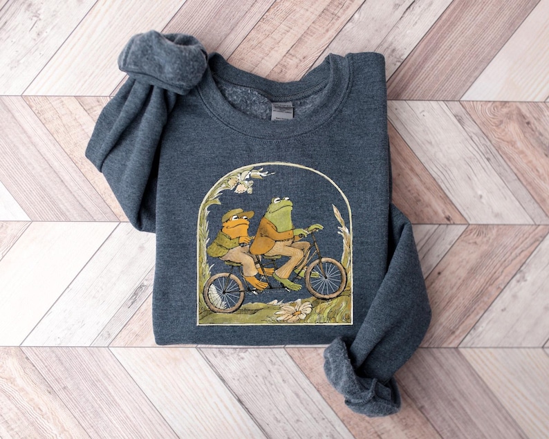 May include: A dark blue sweatshirt with a graphic of two frogs riding a bicycle. The frogs are green and brown and are wearing hats. The bicycle is red and has a basket on the front.