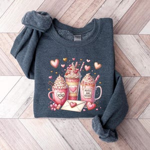 May include: A dark gray sweatshirt with a Valentine's Day design featuring three pink and white hot chocolate mugs with whipped cream and hearts. The mugs have the text "Hugs and Kisses" and "XOXO" on them.
