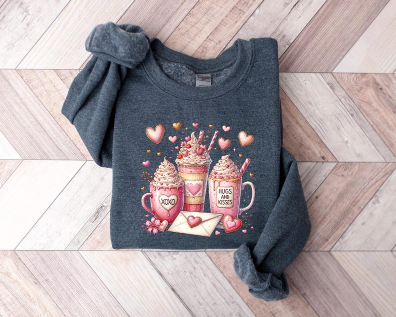 May include: A dark gray sweatshirt with a Valentine's Day design featuring three pink and white hot chocolate mugs with whipped cream and hearts. The mugs have the text "Hugs and Kisses" and "XOXO" on them.