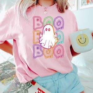Puede incluir: Una camiseta rosa con un fantasma blanco y el texto "BOO" en letras arco&iacute;ris que gotean.