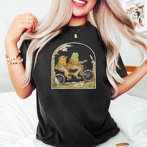 May include: Black t-shirt with a graphic of two frogs riding a bicycle. The frogs are wearing hats and the bicycle has a basket.