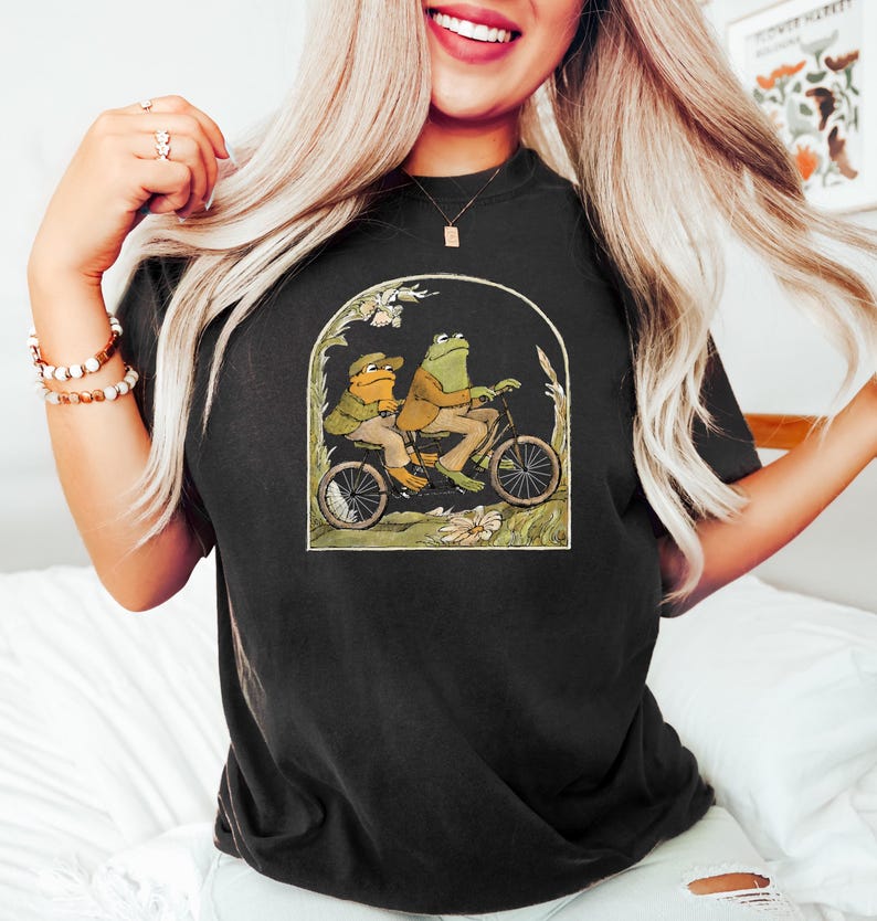May include: Black t-shirt with a graphic of two frogs riding a bicycle. The frogs are wearing hats and the bicycle has a basket.