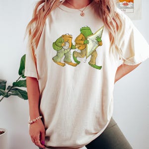 May include: A white t-shirt with a graphic of two frogs. One frog is holding a ball of yarn and the other is holding a kite.