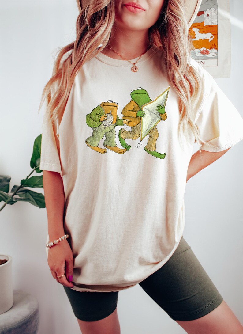 May include: A white t-shirt with a graphic of two frogs. One frog is holding a ball of yarn and the other is holding a kite.