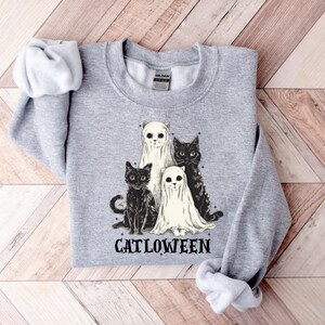 May include: A light gray sweatshirt featuring a graphic of four cats. Two cats are dressed as ghosts, and two are black cats. The word "CATLOWEEN" is printed below the cats in black letters. The sweatshirt is folded on a wooden surface.