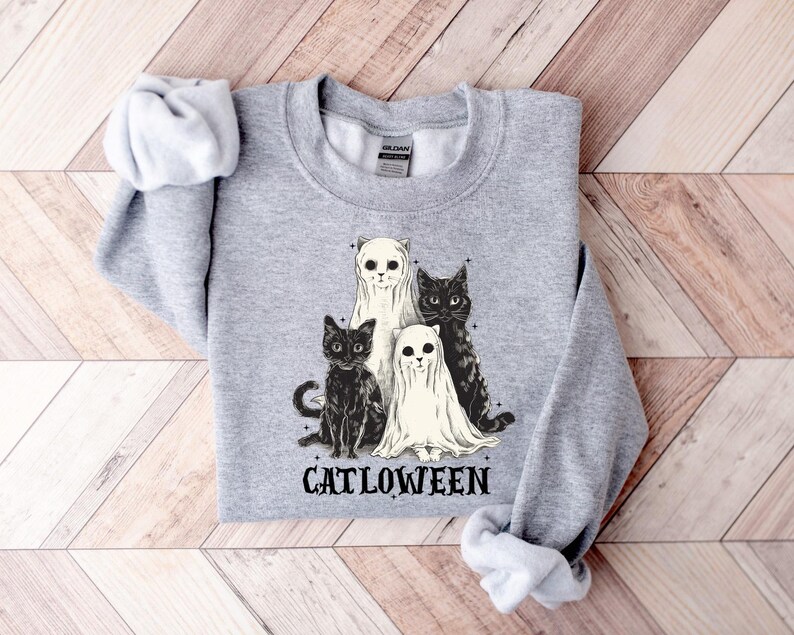 May include: A light gray sweatshirt featuring a graphic of four cats. Two cats are dressed as ghosts, and two are black cats. The word "CATLOWEEN" is printed below the cats in black letters. The sweatshirt is folded on a wooden surface.