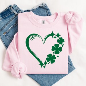 May include: A pink sweatshirt with a green shamrock heart design. The heart is outlined in green and has shamrocks and stars around it.