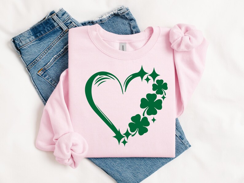 May include: A pink sweatshirt with a green shamrock heart design. The heart is outlined in green and has shamrocks and stars around it.