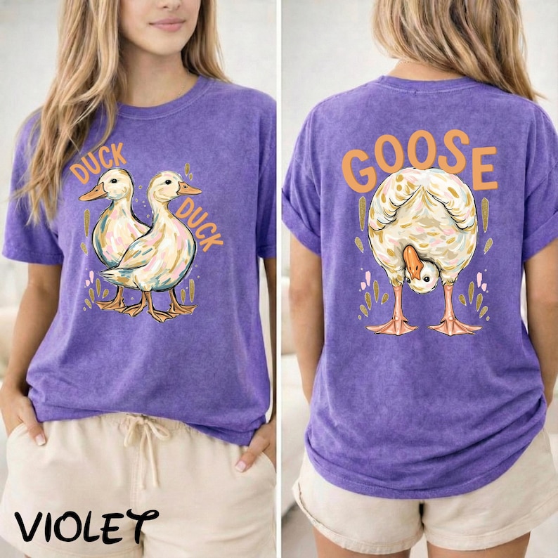 Puede incluir: Camiseta violeta con un gr&aacute;fico de pato en la parte delantera y un gr&aacute;fico de ganso en la parte trasera. La parte delantera de la camiseta tiene la palabra "DUCK" y la parte trasera tiene la palabra "GOOSE" en naranja.