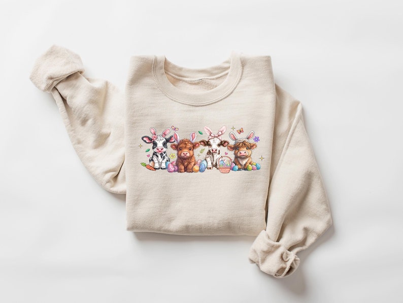 May include: A beige sweatshirt with a graphic of four cows wearing bunny ears and holding Easter eggs. The cows are black and white, brown, and white, and brown and white. The graphic is surrounded by flowers and butterflies.