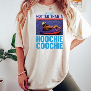 May include: Cream-colored t-shirt with a graphic that reads "It gets hotter than a Hoochie Coochie." The graphic features a person in a cowboy hat floating on a purple inner tube in water.