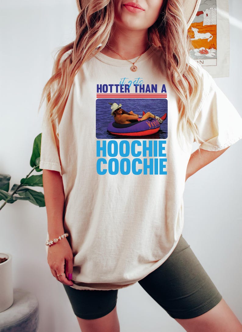 May include: Cream-colored t-shirt with a graphic that reads "It gets hotter than a Hoochie Coochie." The graphic features a person in a cowboy hat floating on a purple inner tube in water.