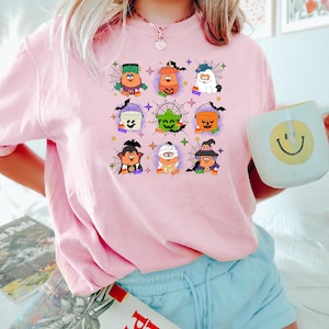 May include: Pink t-shirt with a graphic of nine Halloween characters holding buckets. The characters are dressed as a ghost, a pumpkin, a vampire, a witch, a monster, and a zombie. The graphic is surrounded by spiderwebs and stars.