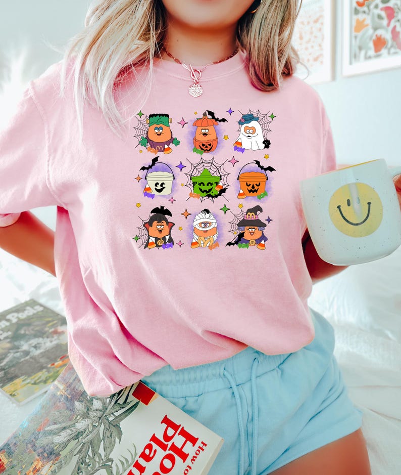 May include: Pink t-shirt with a graphic of nine Halloween characters holding buckets. The characters are dressed as a ghost, a pumpkin, a vampire, a witch, a monster, and a zombie. The graphic is surrounded by spiderwebs and stars.