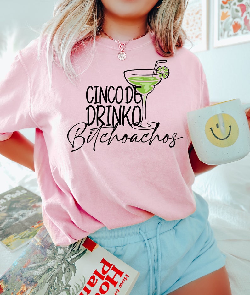 以下が含まれることがあります： ピンクのTシャツに「Cinco de Drinko Bitchoachos」の文字と、ライムのくさび形がついた緑色のマルガリータグラスがプリントされています。