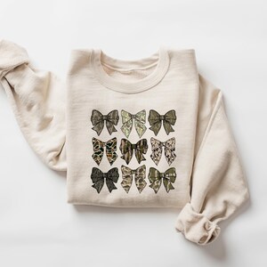 May include: A beige sweatshirt with a design of nine brown and green bows on the front.