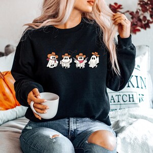 May include: Black sweatshirt featuring a cartoon design of four ghosts dressed as cowboys, complete with hats and bandanas. The ghosts are holding guns and a lasso. The sweatshirt is a crew neck style.
