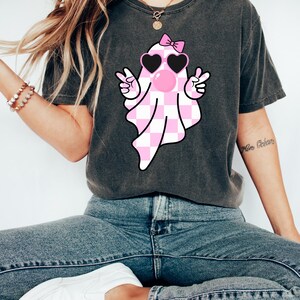 Peut inclure: Un t-shirt gris fonc&eacute; avec un fant&ocirc;me &agrave; carreaux rose et blanc portant des lunettes de soleil et un n&oelig;ud rose. Le fant&ocirc;me fait un signe de paix avec les deux mains et a une bulle rose qui sort de sa bouche.