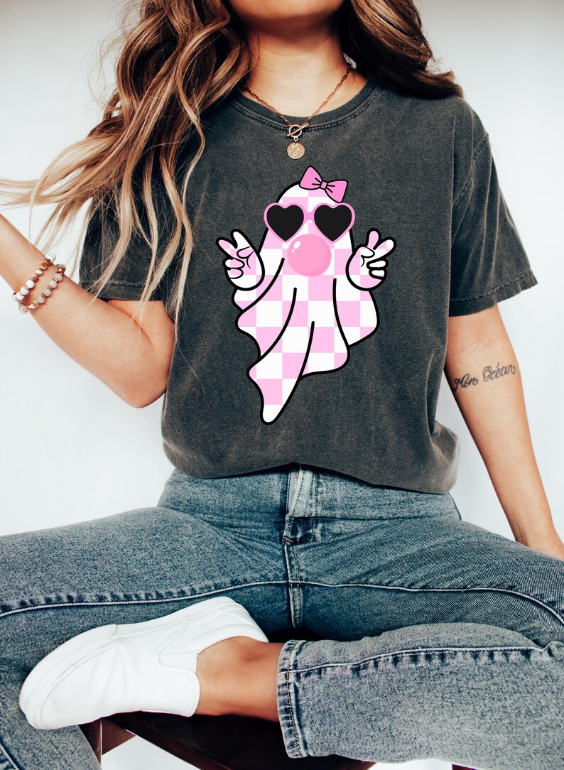 Peut inclure: Un t-shirt gris fonc&eacute; avec un fant&ocirc;me &agrave; carreaux rose et blanc portant des lunettes de soleil et un n&oelig;ud rose. Le fant&ocirc;me fait un signe de paix avec les deux mains et a une bulle rose qui sort de sa bouche.