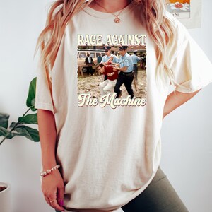 K&ouml;nnte beinhalten: Ein wei&szlig;es T-Shirt mit einer Grafik im Vintage-Stil einer Band namens "Rage Against The Machine". Die Grafik zeigt ein Schwarzwei&szlig;foto eines Mannes, der von zwei Polizisten verhaftet wird.