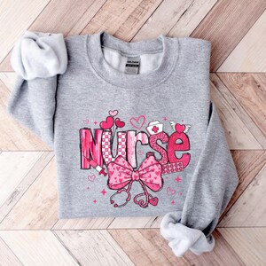 May include: Gray sweatshirt with a pink and white graphic design that says "Nurse" with hearts, a stethoscope, and a bandage.