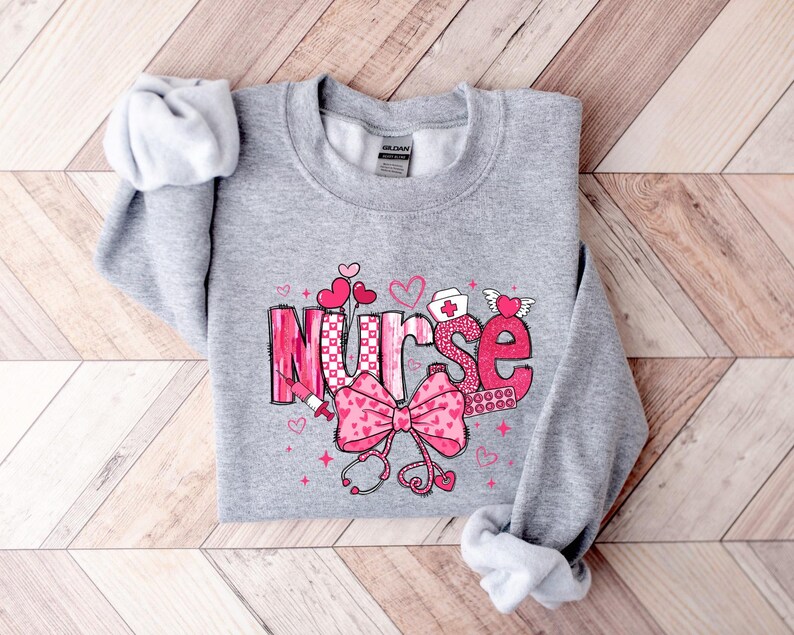 May include: Gray sweatshirt with a pink and white graphic design that says "Nurse" with hearts, a stethoscope, and a bandage.