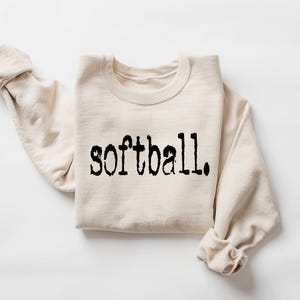 Puede incluir: Una sudadera beige con la palabra "softball" impresa en negro en la parte delantera.
