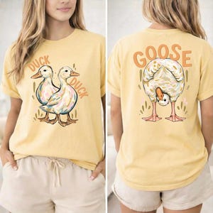 Puede incluir: Camiseta amarilla clara con un dise&ntilde;o de pato en la parte delantera y un dise&ntilde;o de ganso en la parte trasera. La parte delantera presenta dos patos con las palabras "DUCK DUCK". La parte trasera muestra un ganso con la palabra "GOOSE" encima.