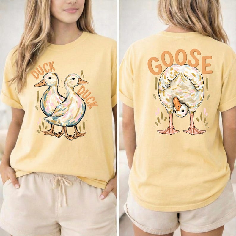 Puede incluir: Camiseta amarilla clara con un dise&ntilde;o de pato en la parte delantera y un dise&ntilde;o de ganso en la parte trasera. La parte delantera presenta dos patos con las palabras "DUCK DUCK". La parte trasera muestra un ganso con la palabra "GOOSE" encima.