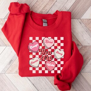 May include: Red sweatshirt with a checkered pattern and hearts that say "You are" with words like "Loved", "Strong", "Capable", "Worthy", and "Enough" inside the hearts.