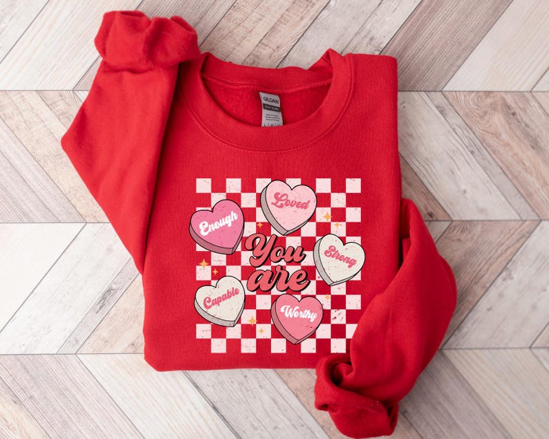 May include: Red sweatshirt with a checkered pattern and hearts that say "You are" with words like "Loved", "Strong", "Capable", "Worthy", and "Enough" inside the hearts.