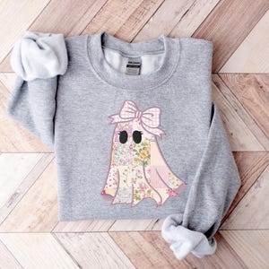 May include: A light gray sweatshirt featuring a ghost design. The ghost has a patchwork body with floral and pink gingham patterns, black eyes, and a pink bow. The sweatshirt is made of a soft material.