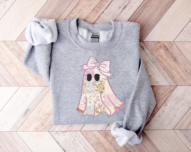 May include: A light gray sweatshirt featuring a ghost design. The ghost has a patchwork body with floral and pink gingham patterns, black eyes, and a pink bow. The sweatshirt is made of a soft material.