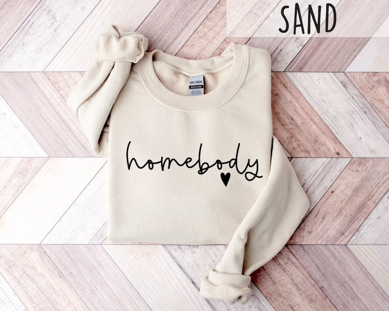 May include: A sand-colored sweatshirt with the text "homebody" in black script with a small black heart.