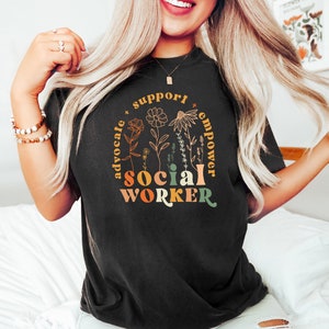 May include: Black t-shirt with a floral design and the text "support advocate social worker empower" in a rainbow color scheme.