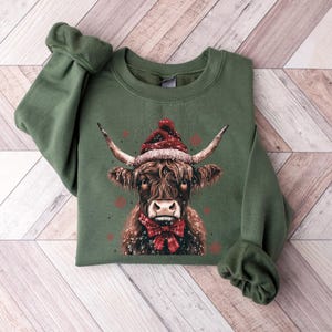 May include: Green sweatshirt with a brown and white highland cow wearing a red Santa hat and a red bow tie. The cow is surrounded by white snowflakes.